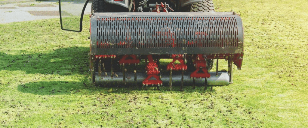 Close-up of a core aeration machine's tine drum pulling cylindrical soil plugs from green turf, with visible aeration holes and loosened soil scattered across the lawn.