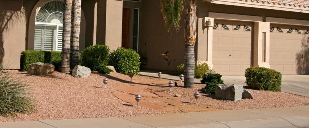 Desert-landscaped front yard with decorative stones, gravel, drought-tolerant shrubs, and palm trees surrounding a stone walkway to a Southwestern-style home.